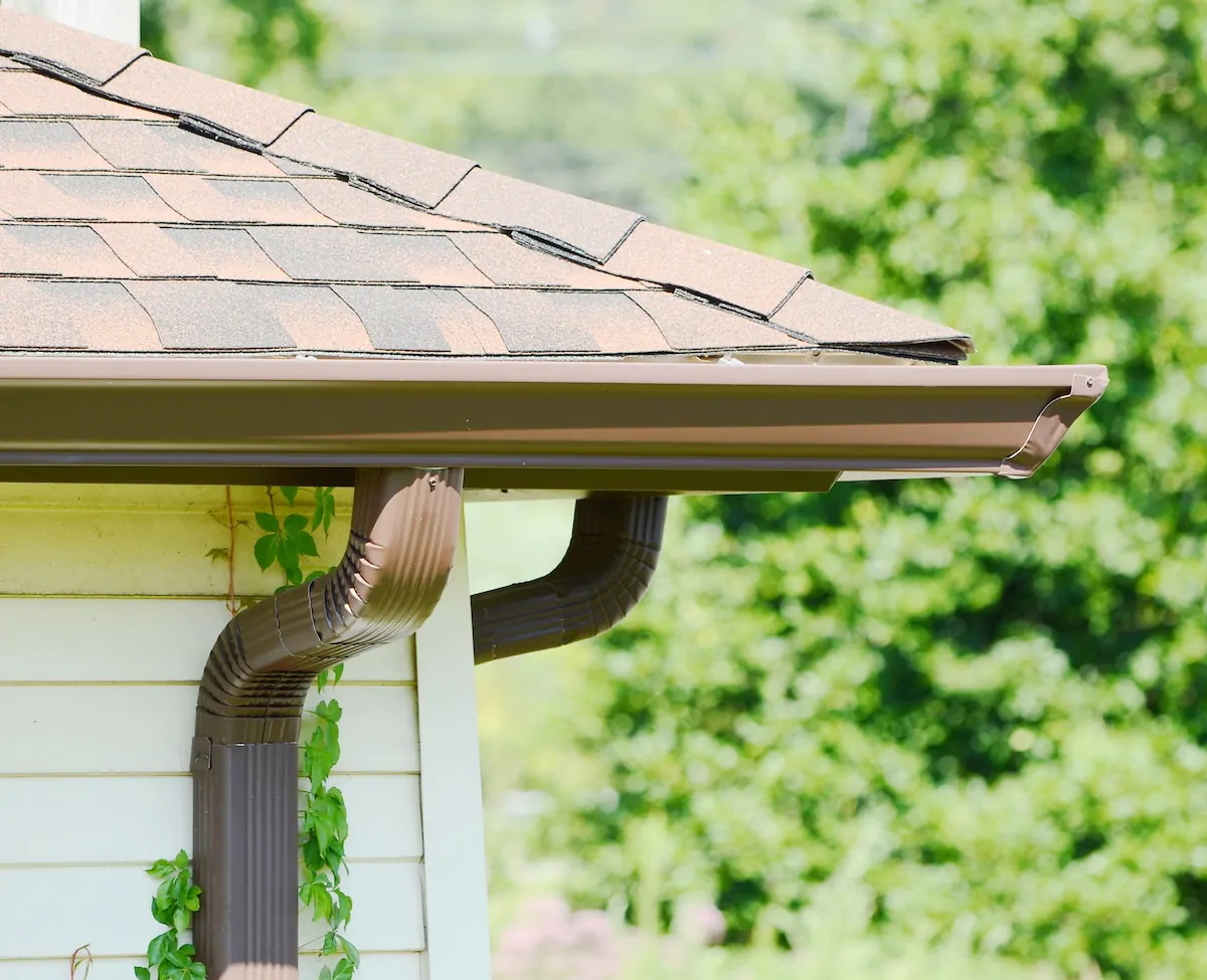 A high-quality professional gutter installation featuring a seamless brown aluminum gutter and matching corrugated downspout with a precise elbow joint, integrated perfectly with a brown asphalt shingle roof.