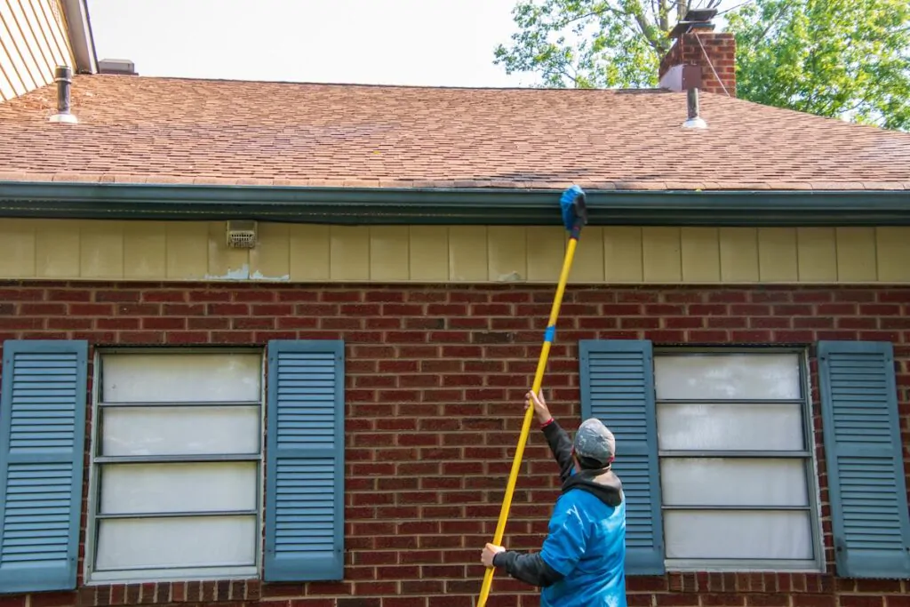 Young man cleaning the soffit of a house with a brush on a long pole. The house has blue shutters and is one story high