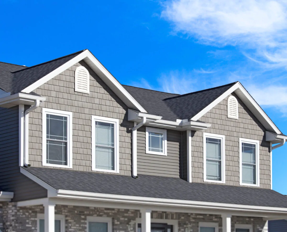 A high-contrast exterior shot of a new home with grey siding and shake-style accents, featuring a dark shingle roof with white ridge vents and matching gutters.