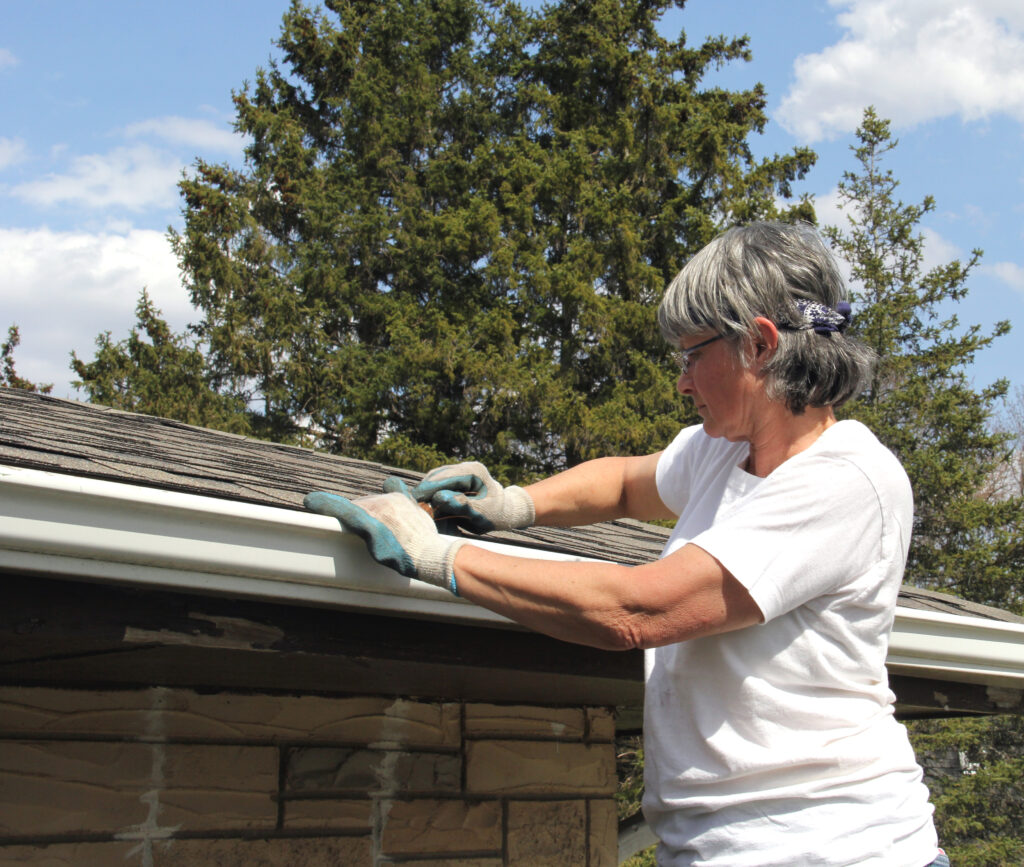 Woman on a ladder cleaning rain gutters with a blue sky in the background.