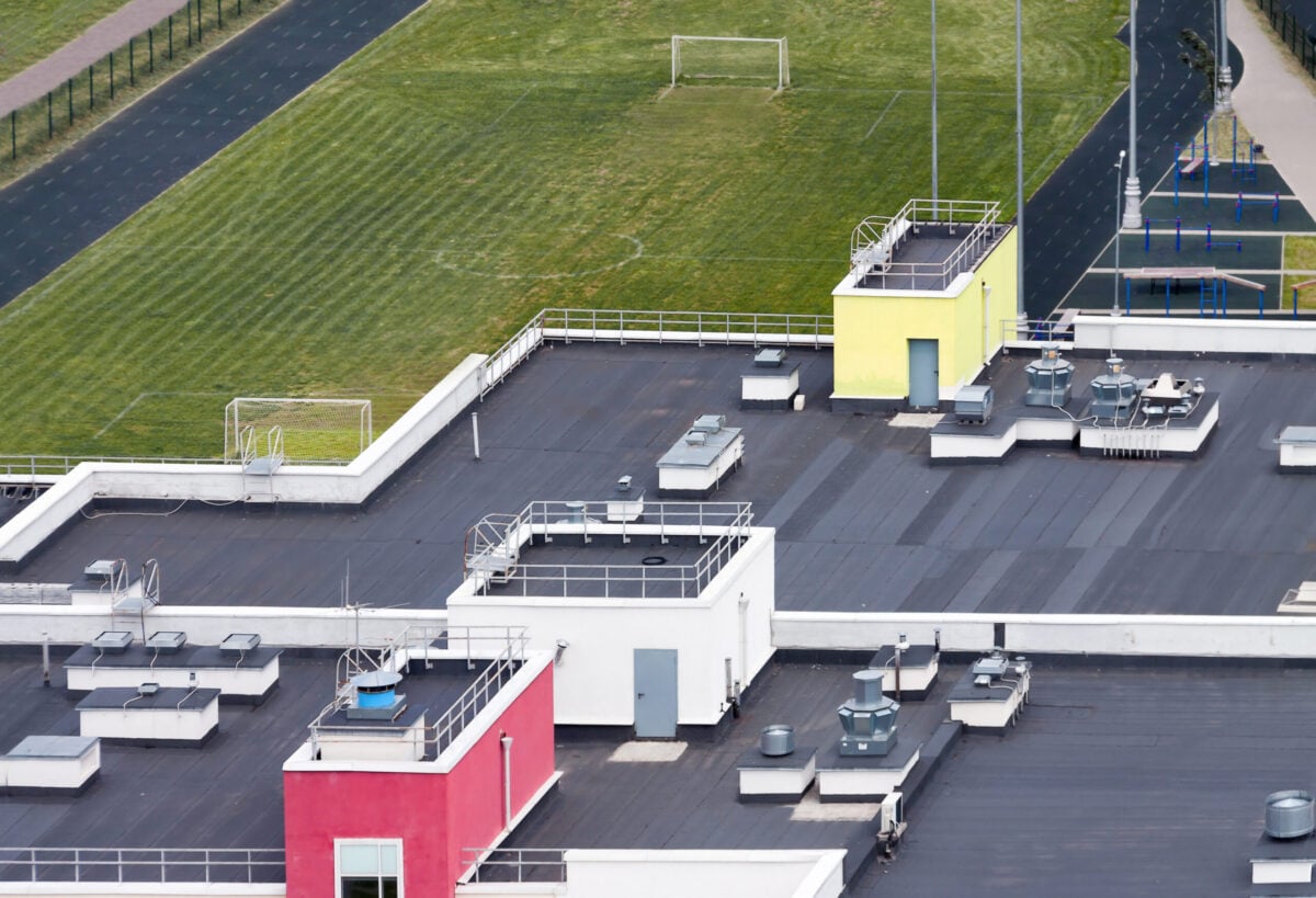 Top view flat roof with air conditioners and hydro insulation membranes on top of a modern apartment building with green football field. Urban development concept - image