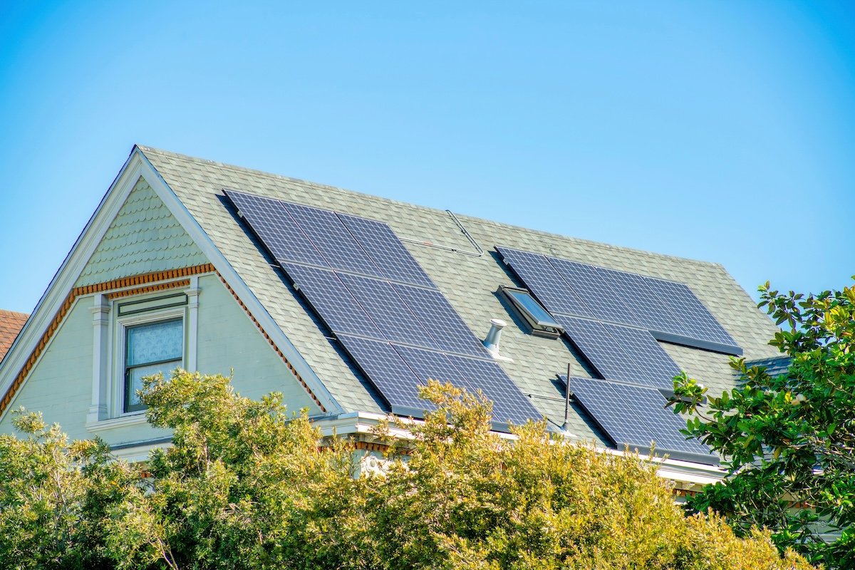 Modern house facade with large solar pannels on roof with light tiles and green stucco with front yard trees and foliage in neighborhood. Blue sky background with no clouds in the city or in suburbs.