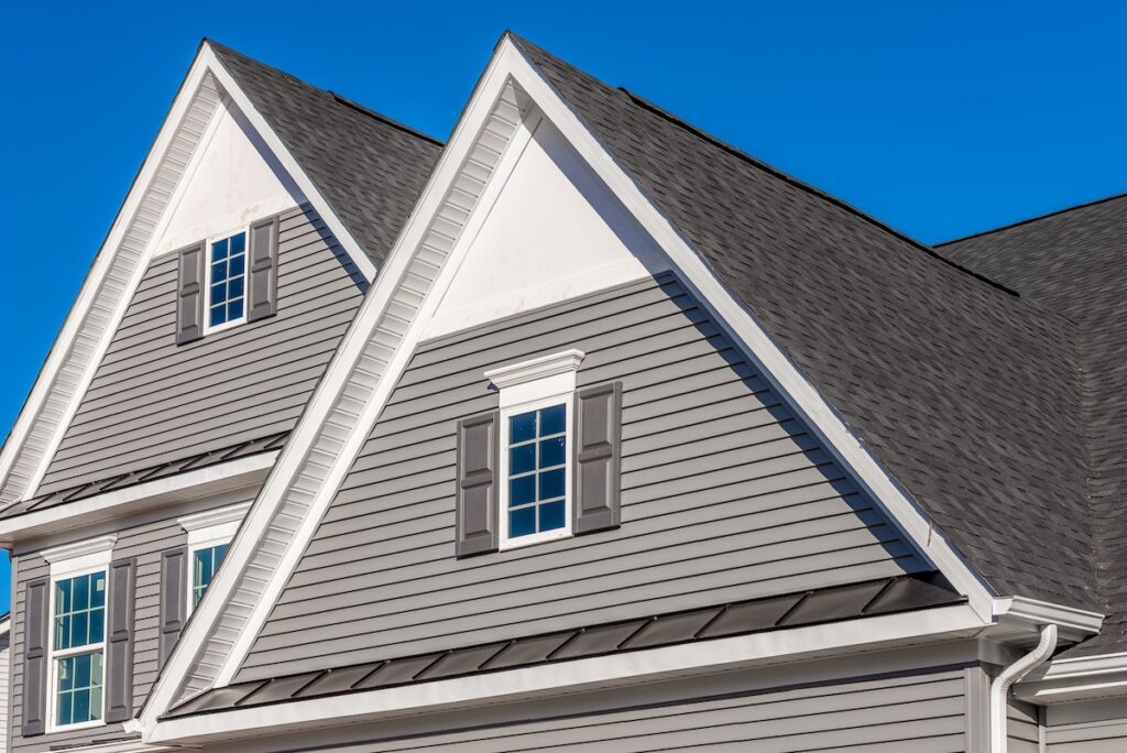 pitched roof Double gable, with white decorative trim over the windows on a triangle gable roof, white soffit and fascia, gray horizontal vinyl lap siding with blue sky background