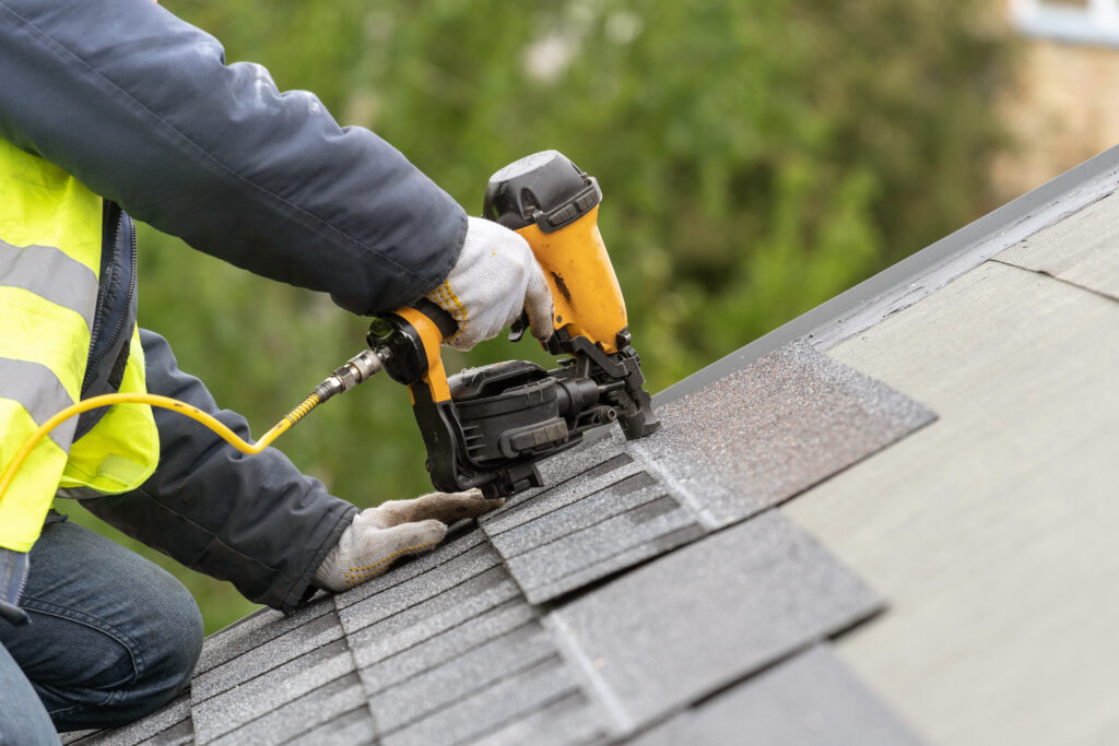 shingle roof repair Unrecognizable roofer worker in uniform work wear using air or pneumatic nail gun and installing asphalt or bitumen tile on top of the roof under construction house