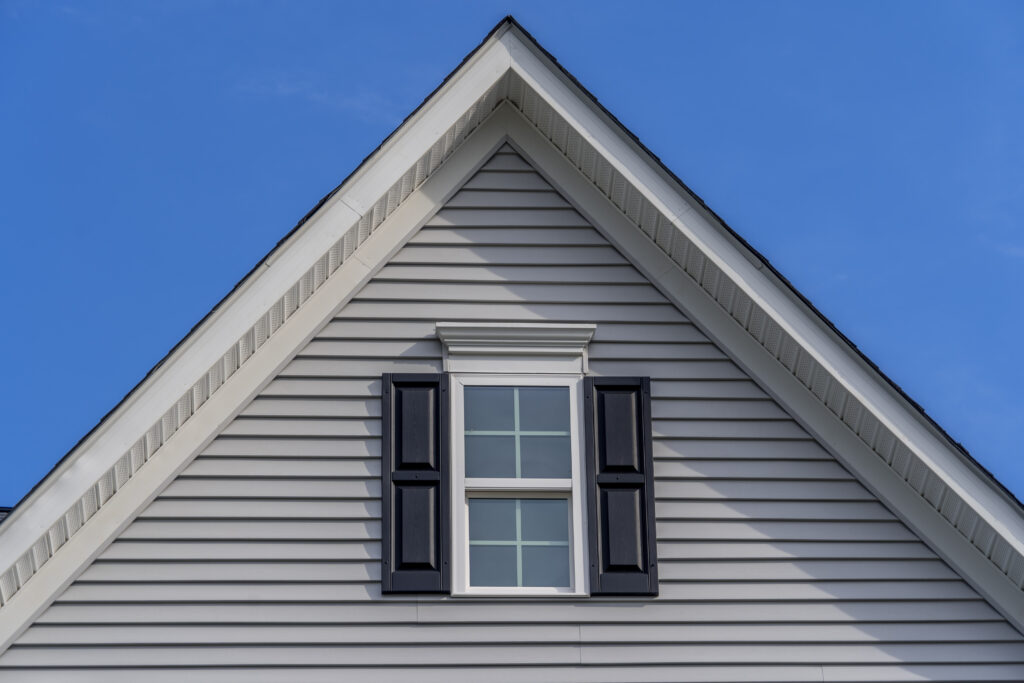 pitched roof Gable with horizontal vinyl lap siding, double hung window with white frame, double vinyl shutters yellow shingle facade on a pitched roof attic at an American single family home neighborhood USA