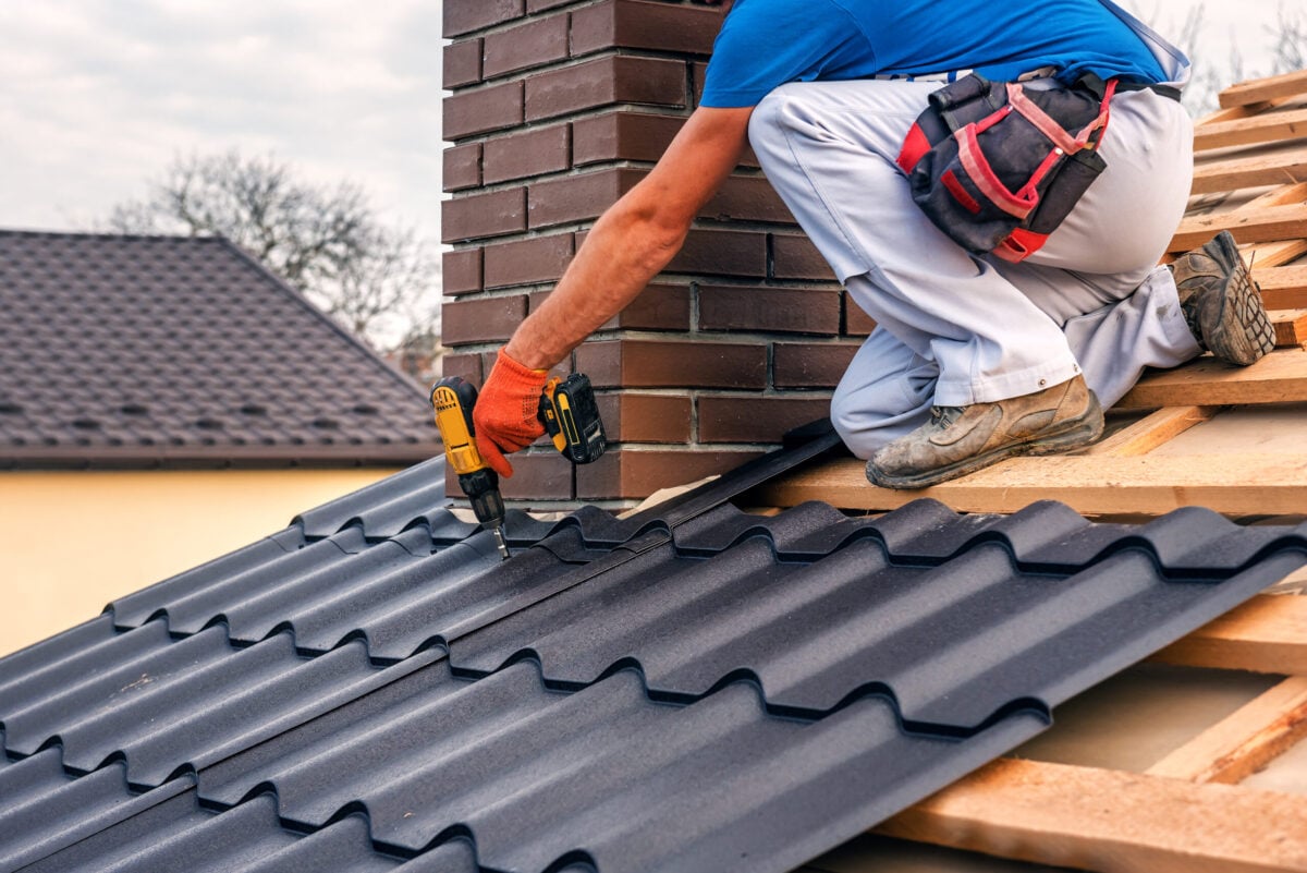 A professional master (roofer) with electric screwdriver covers repairs the roof near the chimney.
