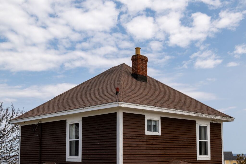 A vintage four sided brown wooden house with an asphalt shingled hip roof.