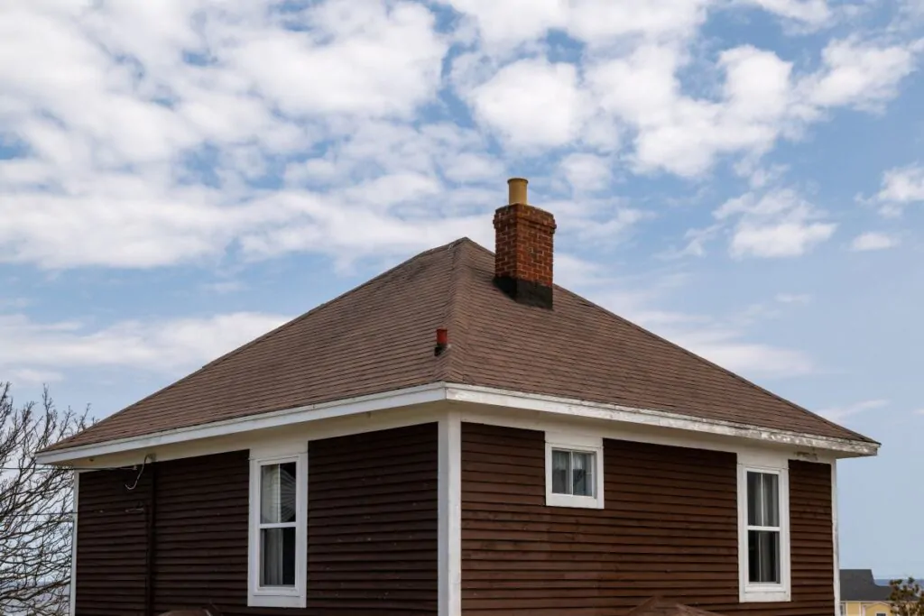 A vintage four sided brown wooden house with an asphalt shingled hip roof.